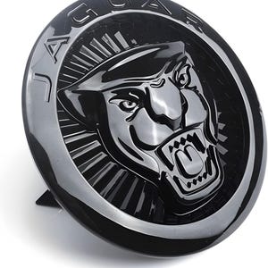 以下が含まれることがあります： 上部にJaguarのブランド名がエンボス加工され、中央に光沢のある黒いジャガーの頭部が浮き彫りにされた円形の黒いエンブレム。ジャガーは口を開けて牙を見せており、背景には放射状のテクスチャが施されている。
