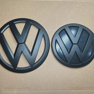Peut inclure: Deux emblèmes Volkswagen circulaires noir mat. L'emblème de gauche présente un design à cadre ouvert avec un logo V et W découpé. L'emblème de droite est une pièce circulaire pleine avec un logo V et W en relief au centre, tous deux en noir.