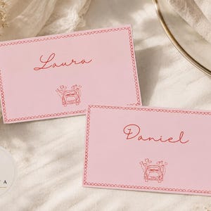 以下が含まれることがあります： 「Laura」と「Daniel」と書かれた赤い筆記体の名前が書かれた2枚のピンク色の席札。各カードには装飾的なボーダーと、ハートの風船が付いた車のイラストが描かれています。カードは白い表面にあります。