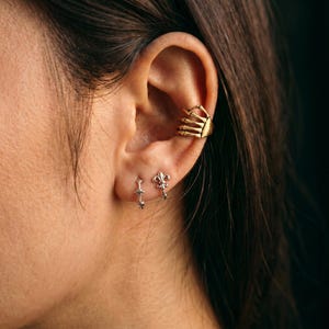 May include: A gold-toned ear cuff shaped like a skeletal hand resting on the cartilage, paired with two silver-toned hoop earrings on the earlobe. One hoop features a small star detail, while the other displays a fleur-de-lis emblem.