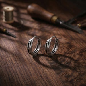 May include: A pair of silver hoop earrings featuring a triple-band design with a dark, oxidized finish. The earrings have a hinged closure mechanism and a rounded, textured profile, showcasing a metallic sheen against a dark wooden surface.