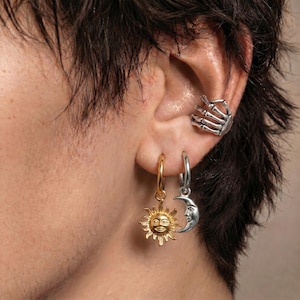Può includere: Un ear cuff argentato a forma di mano scheletrica, abbinato a due orecchini a cerchio. Un cerchio presenta un ciondolo a forma di sole dorato con volto, l'altro un ciondolo a forma di mezzaluna argentata con volto, indossati su un orecchio.