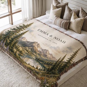 May include: A woven throw blanket featuring a landscape illustration of a mountain range, a lake, and pine trees. The text "EMMA & NOAH" and "SEPTEMBER 19, 2026" is printed on the cream-colored fabric. The blanket has a fringed edge and floral details.