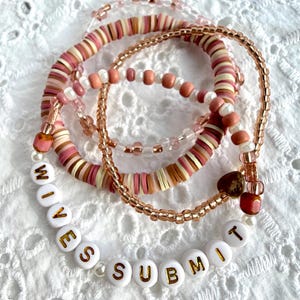 Op de afbeelding: Een stapel van drie kralenarmbanden met platte kleischijfjes in roze, crème en bruine tinten, metallic koperkleurige zaadkralen en ronde witte plastic kralen met gouden letters die de tekst WIVES SUBMIT vormen.