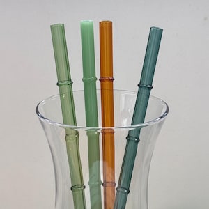 Könnte beinhalten: Vier Glasstrohhalme mit einer strukturierten Bambus-Optik in den Farben Hellgrün, Salbeigrün, Bernstein und Dunkel-Petrol. Die Strohhalme befinden sich in einem klaren, gestielten Glasgefäß mit einem ausgestellten Rand und einem runden Boden.