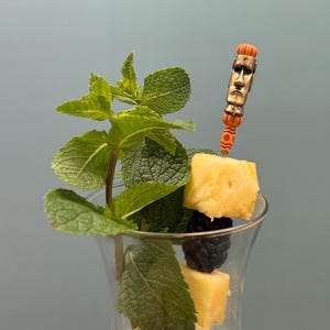 Op de afbeelding: Een helder glas gevuld met gemalen ijs, gegarneerd met een takje verse groene munt, een gedroogde schijfje citrusvrucht en een metalen cocktailprikker. De prikker heeft een oranje en goudkleurig Moai-standbeeldhoofdontwerp met een ananasblokje en een braam.