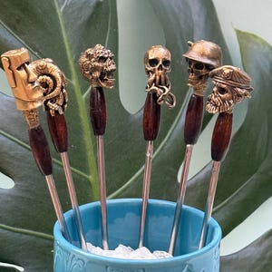 Könnte beinhalten: Ein Set aus sechs Cocktailspießen aus Metall mit dunkelbraunen Holzgriffen und detaillierten bronzefarbenen Aufsätzen, darunter eine Moai-Statue, ein Totenkopf mit Tentakeln, ein behelmter Totenkopf und ein bärtiger Seemann in einem hellblauen Becher.
