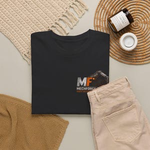 May include: A black t-shirt with the text "MF MECHFORGE ROBOTIC WELDING" and a graphic of a robotic arm. The shirt is laid flat with beige pants, a woven mat, and a candle in the background.