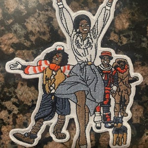 May include: Embroidered patch featuring a group of figures in a celebratory pose. The central figure, a woman, has her arms raised. Other figures include a person in a striped outfit, and a person in a red and black outfit. A small dog is also present.