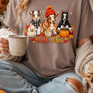 May include: A pebble-colored t-shirt featuring a graphic of three goats, one wearing a black bandana and one wearing an orange beanie, surrounded by pumpkins, autumn leaves, and a coffee mug, with the text Fall Vibes printed in orange and red.