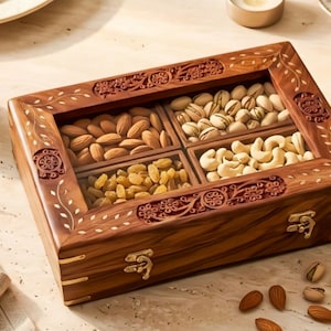 Op de afbeelding: Een rechthoekige houten doos met een glazen deksel, voorzien van gedetailleerd bloemmotiefsnijwerk en messing sluitingen. De binnenkant is verdeeld in vier vakken met amandelen, pistachenoten, cashewnoten en rozijnen, met enkele losse noten.