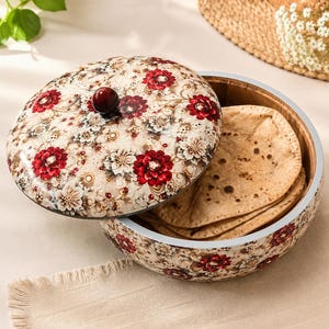 Peut inclure: Un récipient rond en bois avec couvercle, orné d'un motif floral de fleurs rouge foncé et blanches sur fond crème. Le couvercle possède un bouton marron foncé. À l'intérieur se trouvent plusieurs pains plats ronds, fins et brun clair.