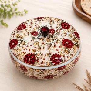 Puede incluir: Un recipiente redondo de cerámica con tapa que presenta un patrón floral detallado con flores de color rojo intenso y crema, detalles dorados intrincados y pequeños puntos rojos. La tapa tiene un pequeño pomo esférico de color rojo oscuro.