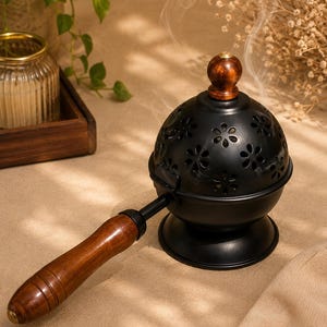 May include: A black metal incense burner with a dome-shaped lid featuring cut-out flower patterns and a small wooden knob. The burner has a long, polished wooden handle and a sturdy circular base, with thin wisps of smoke rising from the top.
