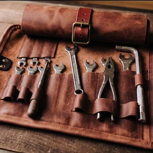 Può includere: Un rotolo di attrezzi in pelle marrone con vari utensili a mano, tra cui chiavi inglesi, pinze e un cacciavite. Il rotolo è fissato con un cinturino e una fibbia in pelle. Gli attrezzi sono ordinatamente organizzati in tasche individuali.