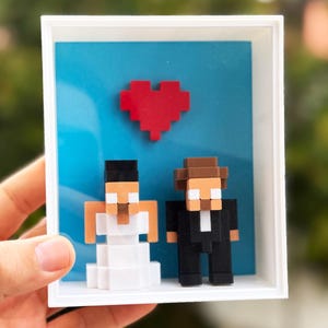 May include: A framed pixel art wedding scene. The artwork features a red heart above two blocky figures, a bride in white and a groom in a black suit, set against a blue background. The frame is white.
