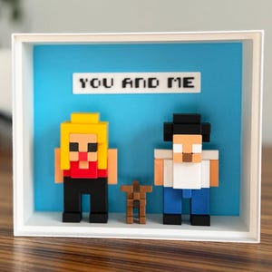 May include: A framed pixel art piece with two figures and a small table. The figures are in a boxy style, with one blonde and one with black hair. The background is blue, and the text "YOU AND ME" is above the figures.