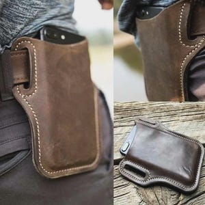 May include: A brown leather phone holster featuring visible white stitching and integrated belt loops. The holster is designed to hold a smartphone securely and is shown both attached to a belt and as a separate item on a wooden surface.