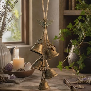 May include: A hanging wind chime featuring five antique brass-toned metal bells suspended by jute twine. The top of the arrangement is adorned with a metal bee ornament, while the bells are accented with small metal charms including a key and a bee.