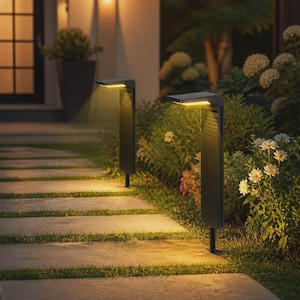 Puede incluir: Dos luces de camino exteriores negras y rectangulares con paneles LED integrados, montadas sobre estacas delgadas. Las luces emiten un brillo cálido hacia abajo que ilumina el camino de piedra y el follaje verde circundante en un jardín.