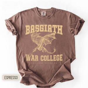 以下が含まれることがあります： エスプレッソ色のTシャツにドラゴンのグラフィック。シャツには、大学風フォントで「BASGIATH WAR COLLEGE」の文字と、その下に「DRAGON RIDERS」の文字がプリントされています。ドラゴンは翼を広げた姿で描かれています。