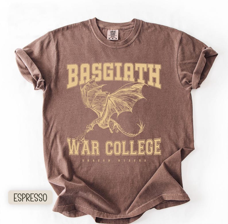 以下が含まれることがあります： エスプレッソ色のTシャツにドラゴンのグラフィック。シャツには、大学風フォントで「BASGIATH WAR COLLEGE」の文字と、その下に「DRAGON RIDERS」の文字がプリントされています。ドラゴンは翼を広げた姿で描かれています。