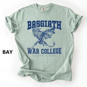 以下が含まれることがあります： 水色のTシャツ。青色の文字で「BASGIATH WAR COLLEGE」と書かれており、ドラゴンのイラストが描かれています。左側には「BAY」の文字も表示されています。