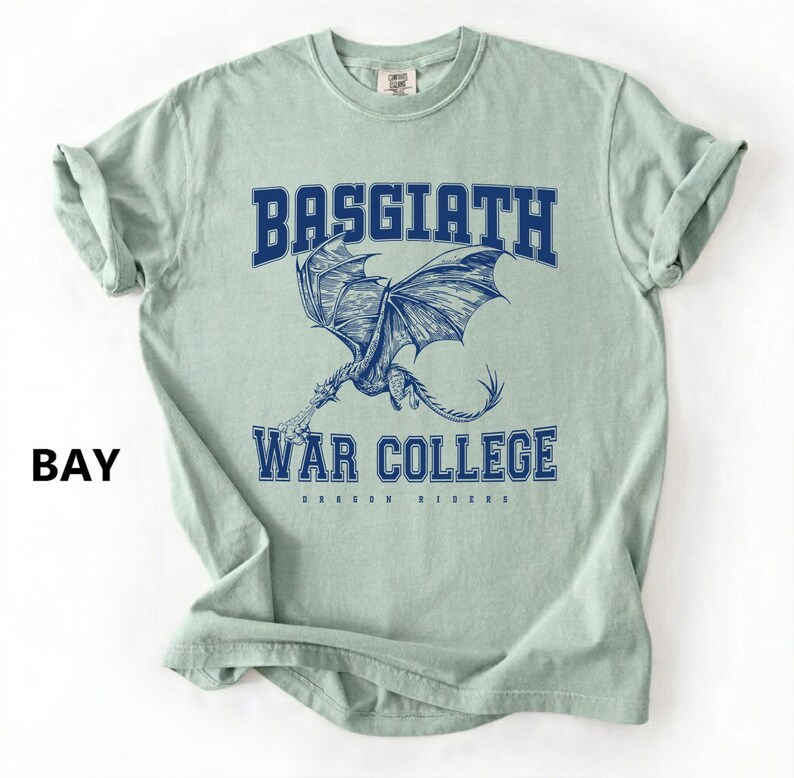 以下が含まれることがあります： 水色のTシャツ。青色の文字で「BASGIATH WAR COLLEGE」と書かれており、ドラゴンのイラストが描かれています。左側には「BAY」の文字も表示されています。