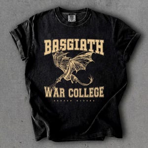 以下が含まれることがあります： 黒色のTシャツ。カレッジフォントで「BASGIATH WAR COLLEGE」の文字がプリントされています。 「BASGIATH」の下にはドラゴンの詳細なイラストが描かれています。「WAR COLLEGE」の下には「DRAGON RIDERS」の文字がプリントされています。