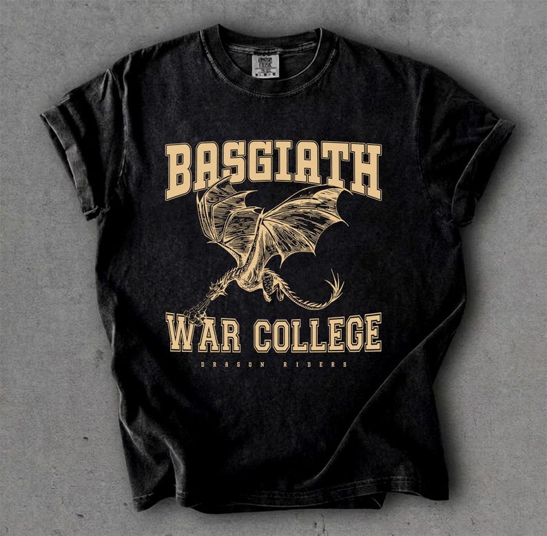 以下が含まれることがあります： 黒色のTシャツ。カレッジフォントで「BASGIATH WAR COLLEGE」の文字がプリントされています。 「BASGIATH」の下にはドラゴンの詳細なイラストが描かれています。「WAR COLLEGE」の下には「DRAGON RIDERS」の文字がプリントされています。