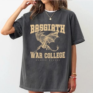 以下が含まれることがあります： 濃いグレーのTシャツ。ベージュの大学風フォントで「BASGIATH WAR COLLEGE」と書かれています。翼を広げたドラゴンの詳細なイラストがテキストの下に配置されています。「DRAGON RIDERS」の文字が下にあります。
