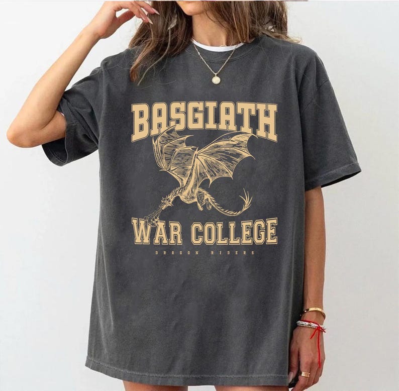 以下が含まれることがあります： 濃いグレーのTシャツ。ベージュの大学風フォントで「BASGIATH WAR COLLEGE」と書かれています。翼を広げたドラゴンの詳細なイラストがテキストの下に配置されています。「DRAGON RIDERS」の文字が下にあります。