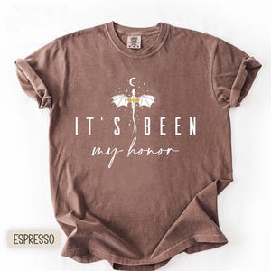 Puede incluir: Camiseta color espresso con el texto "IT'S BEEN my honor" en escritura blanca. Un gr&aacute;fico de drag&oacute;n blanco con detalles dorados est&aacute; centrado sobre el texto, con una luna creciente y estrellas. La camiseta est&aacute; doblada.