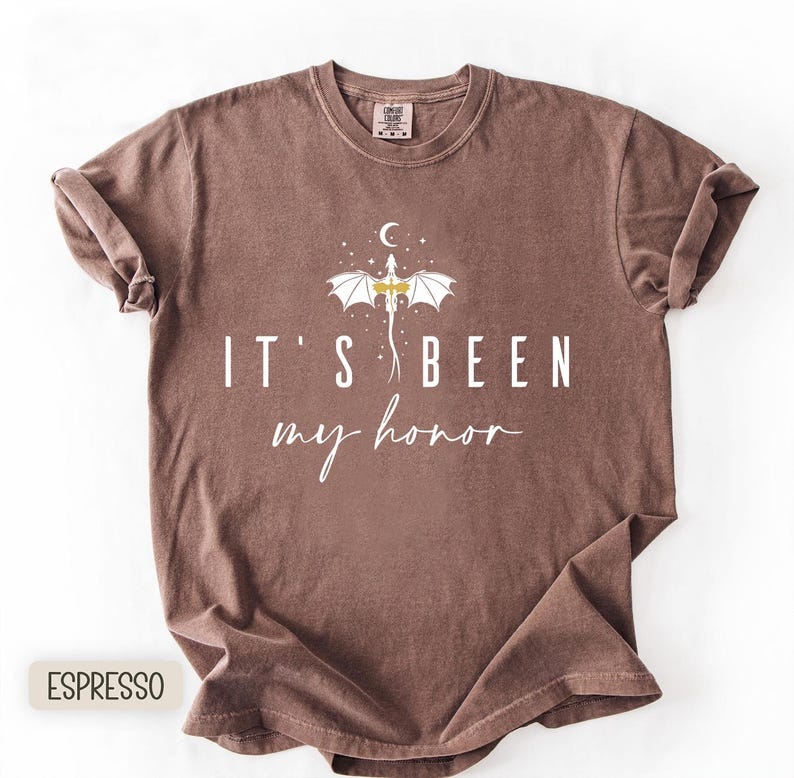 Puede incluir: Camiseta color espresso con el texto "IT'S BEEN my honor" en escritura blanca. Un gr&aacute;fico de drag&oacute;n blanco con detalles dorados est&aacute; centrado sobre el texto, con una luna creciente y estrellas. La camiseta est&aacute; doblada.