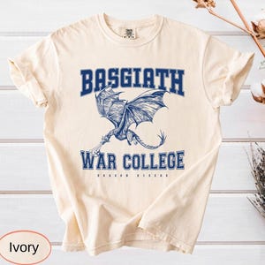 以下が含まれることがあります： アイボリー色のTシャツ。青いドラゴンのグラフィックと「BASGIATH WAR COLLEGE」の文字が描かれています。「DRAGON RIDERS」の文字は「WAR COLLEGE」の下にあります。様々な服装に合うニュートラルカラーの衣服です。