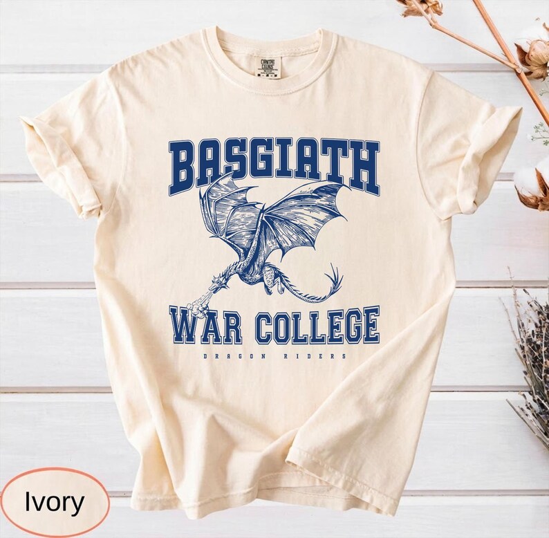 以下が含まれることがあります： アイボリー色のTシャツ。青いドラゴンのグラフィックと「BASGIATH WAR COLLEGE」の文字が描かれています。「DRAGON RIDERS」の文字は「WAR COLLEGE」の下にあります。様々な服装に合うニュートラルカラーの衣服です。