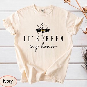 Puede incluir: Camiseta de color marfil con el texto negro "IT'S BEEN my honor". Un dise&ntilde;o de drag&oacute;n negro con detalles dorados se encuentra sobre el texto. La camiseta tiene cuello redondo y mangas dobladas.