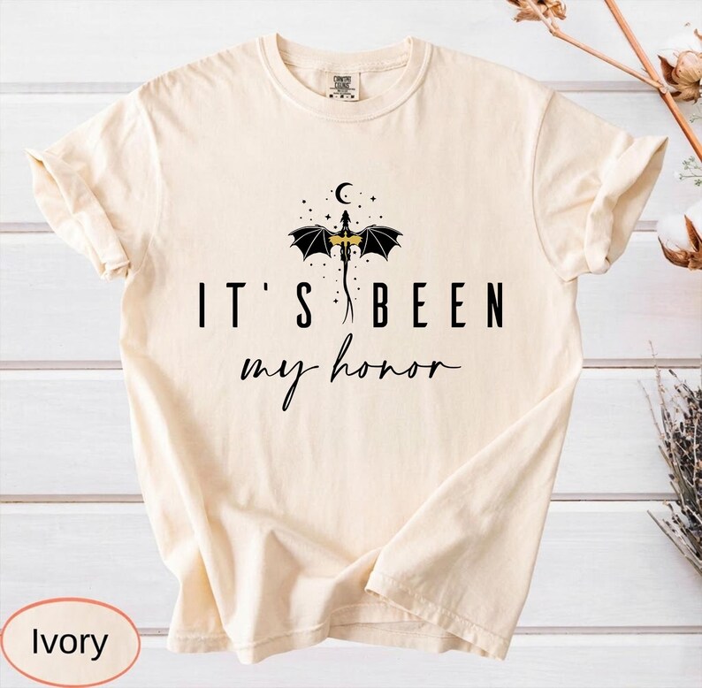 Puede incluir: Camiseta de color marfil con el texto negro "IT'S BEEN my honor". Un dise&ntilde;o de drag&oacute;n negro con detalles dorados se encuentra sobre el texto. La camiseta tiene cuello redondo y mangas dobladas.