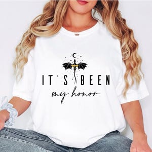 Puede incluir: Camiseta blanca con el texto negro "IT'S BEEN my honor". Encima del texto hay un drag&oacute;n negro con cuerpo dorado, alas y una luna creciente y estrellas. La camiseta es de estilo casual.