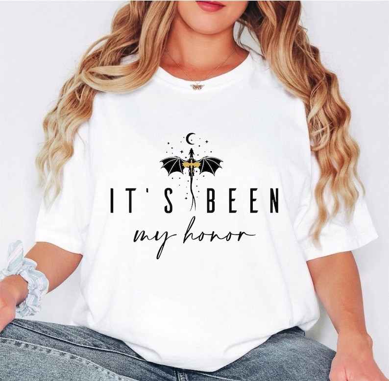 Puede incluir: Camiseta blanca con el texto negro "IT'S BEEN my honor". Encima del texto hay un drag&oacute;n negro con cuerpo dorado, alas y una luna creciente y estrellas. La camiseta es de estilo casual.