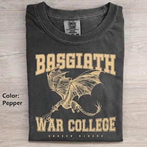 以下が含まれることがあります： ドラゴンが描かれたペッパーカラーのTシャツ。「BASGIATH」と「WAR COLLEGE」の文字がカレッジフォントでプリントされています。Tシャツには「DRAGON RIDERS」の文字も含まれています。Tシャツは折りたたまれています。