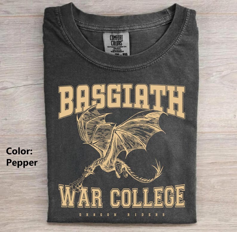 以下が含まれることがあります： ドラゴンが描かれたペッパーカラーのTシャツ。「BASGIATH」と「WAR COLLEGE」の文字がカレッジフォントでプリントされています。Tシャツには「DRAGON RIDERS」の文字も含まれています。Tシャツは折りたたまれています。