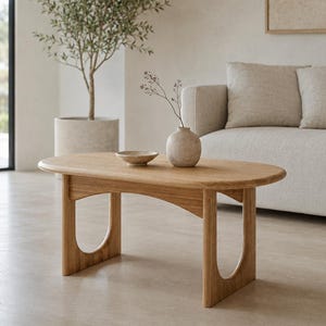 Puede incluir: Una mesa de centro ovalada de madera con acabado marrón claro, que cuenta con cuatro patas conectadas por soportes arqueados. La superficie de la mesa sostiene un pequeño cuenco de cerámica redondo y claro y un jarrón esférico con una rama seca.