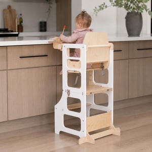 Può includere: Una torre di apprendimento convertibile in seggiolone in legno con finitura in legno chiaro naturale e pannelli laterali bianchi. La struttura include una seduta con schienale, un vassoio e una base robusta con barre di supporto orizzontali.