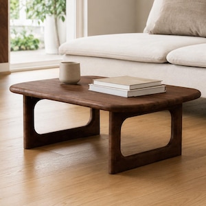 Puede incluir: Una mesa de centro de madera de color marrón oscuro con esquinas redondeadas y una base rectangular abierta. Una pequeña taza de cerámica de color claro y dos libros apilados con cubiertas lisas están colocados sobre la superficie plana.