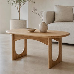 Peut inclure: Une table basse ovale en bois avec une finition brun clair, dotée de quatre pieds en forme de U. Sur le plateau se trouvent un petit bol en céramique ronde de couleur claire et un vase assorti contenant une tige botanique séchée.
