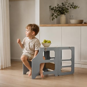 May include: A light gray wooden learning tower with a convertible design, featuring cut-out side panels and metal latch hardware. The structure includes a platform for standing or sitting, finished in a smooth matte gray paint with visible wood grain.