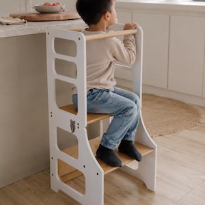 May include: A white wooden kitchen helper stool with a natural wood finish platform and steps. The structure features side panels with cut-out openings, a horizontal top support bar, and a metal latch mechanism on the side for folding functionality.