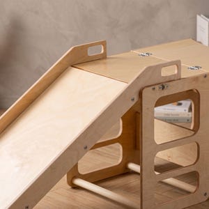 May include: A light-coloured wooden climbing structure featuring a smooth, angled slide attached to a platform. The unit includes side panels with cut-out handles, horizontal support bars, and metal hinges connecting the flat top sections.