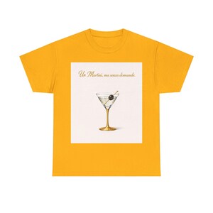 Può includere: T-shirt giallo senape con grafica di un martini in un bicchiere dorato con un'oliva. La grafica è su sfondo bianco con la scritta "Un Martini, ma senza domande."